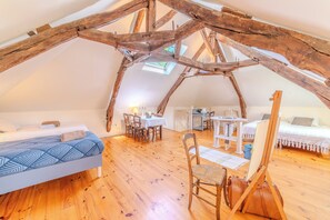 A loft-style room with exposed wooden beams, a bed, a dining table with chairs, and an easel.