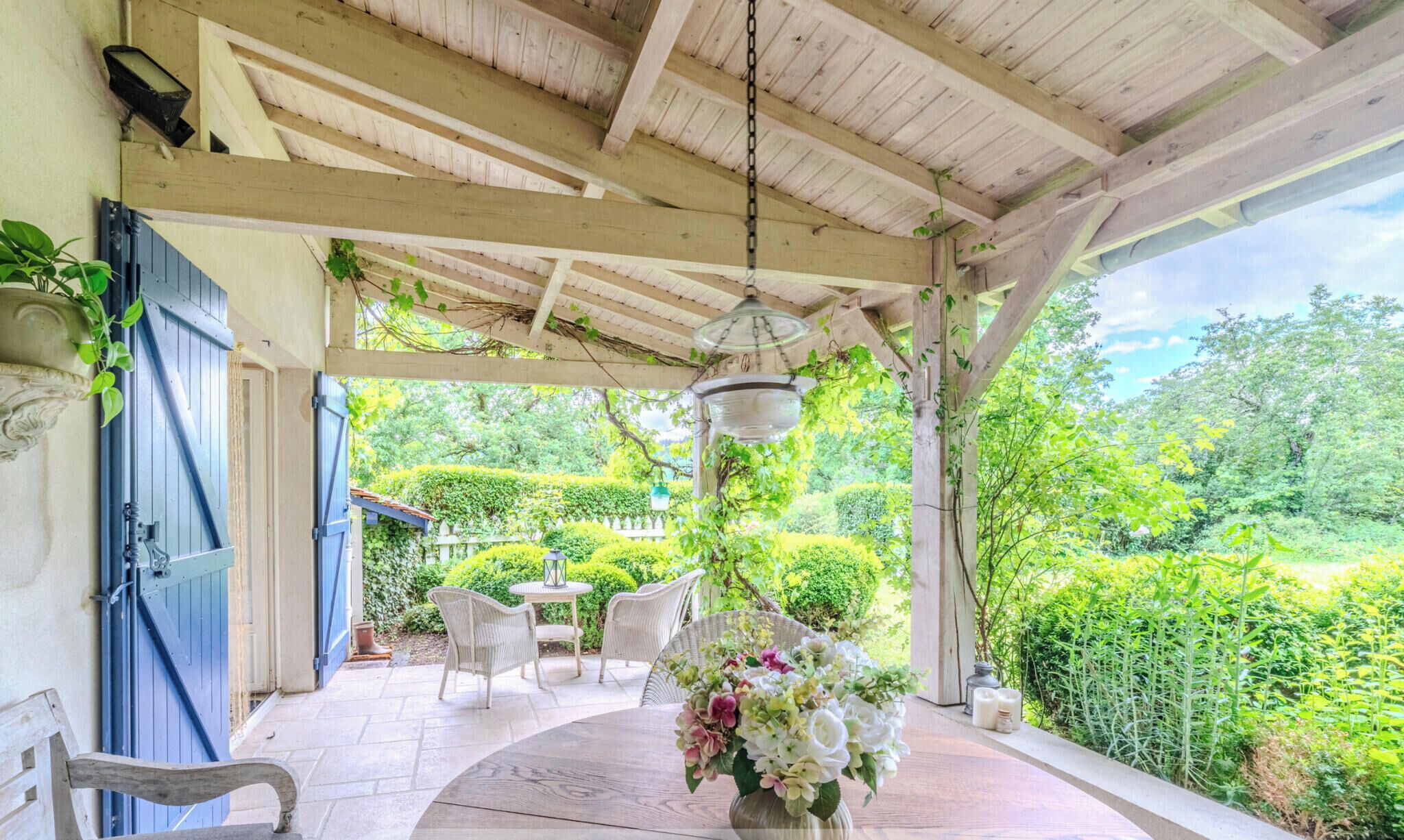 A covered patio with a table and chairs, a hanging light fixture, and a view of a lush garden.