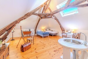 A cozy attic bedroom with a sloped ceiling, a wooden beam structure, a bed, a desk, a chair, a small table, a lamp, and a window.