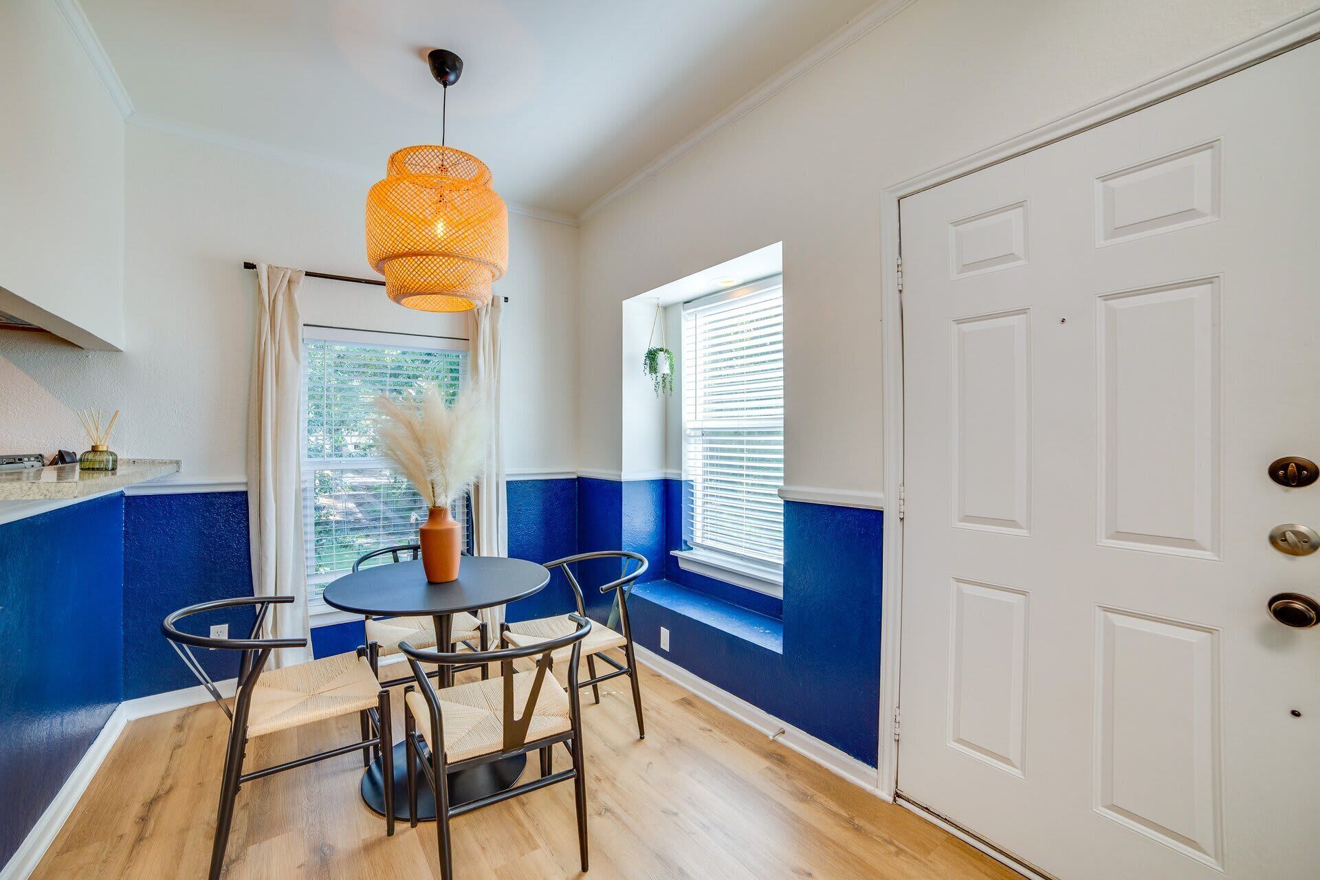 A room with a round table, chairs, a hanging light fixture, a window with blinds, and a door.