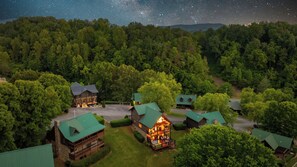 Eine Lodge mit mehreren Holzhäuschen, einem zentralen Gebäude mit beleuchteter Veranda, umgeben von dichtem Wald und sternenklarer Nacht.