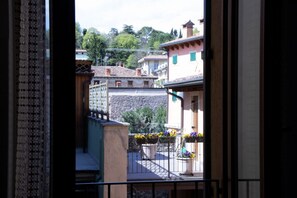 Blick durch ein Fenster mit gemustertem Vorhang, ein Balkon mit Pflanzgefäßen und Gebäude mit traditioneller Architektur im Hintergrund.