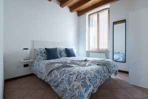 Ein Schlafzimmer mit Bett, Nachttisch, Fenster mit Jalousien und Spiegel an der Wand.