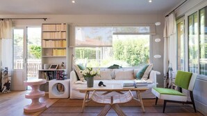 Ein modernes Wohnzimmer mit einer Couch, einem Couchtisch, einem Sessel, einem Bücherregal und einem Fenster mit Blick ins Grüne.