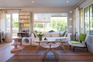 Ein modernes Wohnzimmer mit einer Couch, einem Couchtisch, einem Sessel, einem Bücherregal und einem Fenster mit Blick ins Grüne.