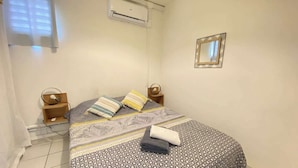 A bedroom with a bed, two bedside tables, an air conditioning unit, a mirror, and a window with blinds.