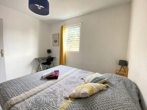 A neatly made bed with a patterned bedspread and pillows, a desk with a chair, a small table with a lamp, and a window with curtains.