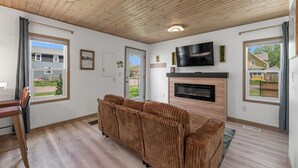Ett vardagsrum med ett träpanelat tak, en tv som är monterad ovanför en öppen spis, en brun soffa och ett matbord med stolar.