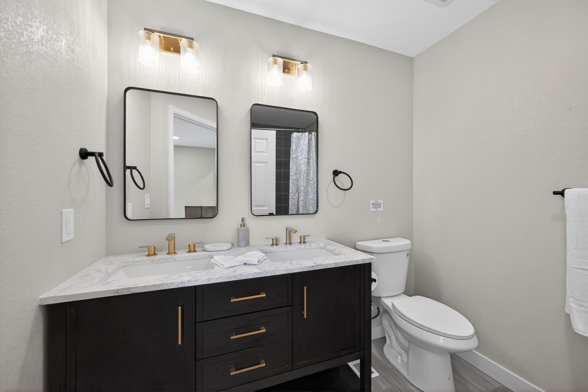 A modern bathroom with a marble sink, dark cabinets, a toilet, and two wall-mounted mirrors with light fixtures.