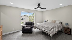 Ein Schlafzimmer mit einem Bett, einem Sessel, einem Nachttisch, einer Lampe, einem Deckenventilator und einem Fenster.
