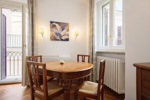 Ein Esszimmer mit einem runden Holztisch, vier Stühlen, zwei Wandleuchten, einem Gemälde und einem Fenster mit Blick auf einen Balkon.