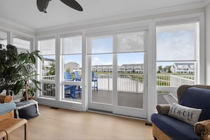 Una habitación con ventanas amplias, vista a una zona residencial, una silla de mimbre y un cojín azul con el texto 'BEACH'.