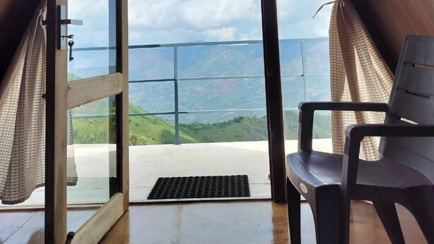 A room with a large glass door leading to a balcony with a view of green hills. - Adventure trails (Kodaikanal)