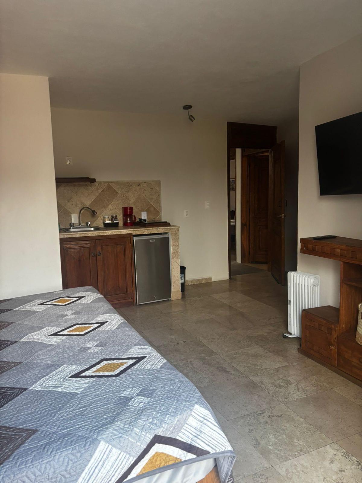 A bedroom with a bed, a kitchenette with a sink and a coffee maker, a hallway leading to a door, and a radiator.