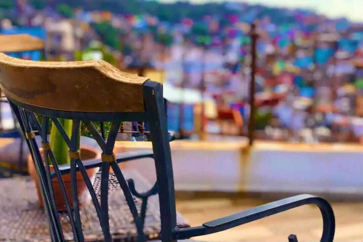 A chair with a wooden backrest and metal frame, positioned outdoors with a blurred background of buildings and a body of water.