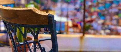 Una silla con respaldo de madera y estructura metálica, ubicada al aire libre con un fondo borroso de edificios y una masa de agua.