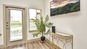 Una habitación con puerta, ventana, planta en maceta, una mesa consola dorada y televisor montado en la pared.