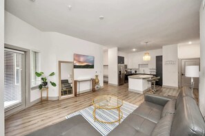 A modern living room with a sofa, a round coffee table, a TV, and a kitchen area in the background. - Modern Houston Escape Bowling, Pool Table & Gym (Houston)
