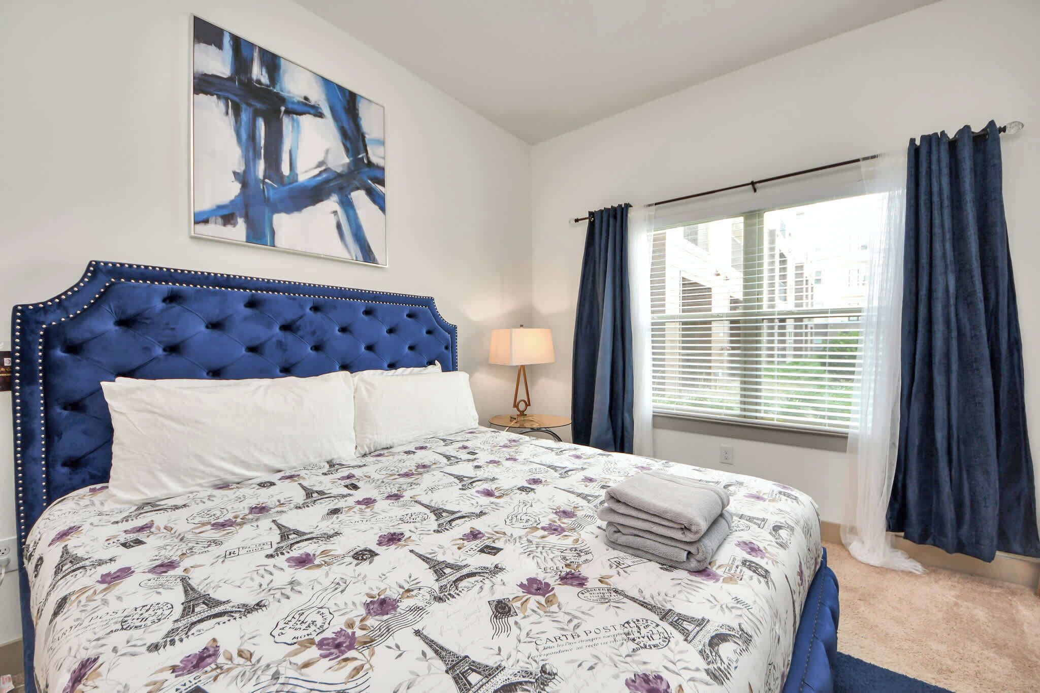 A bedroom with a blue tufted headboard, a bed with a Paris-themed bedspread, a nightstand with a lamp, and a window with curtains.