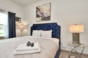 Habitación de hotel con una cama grande, cabecero, dos lámparas de noche, una mesita de vidrio y un cuadro en la pared.