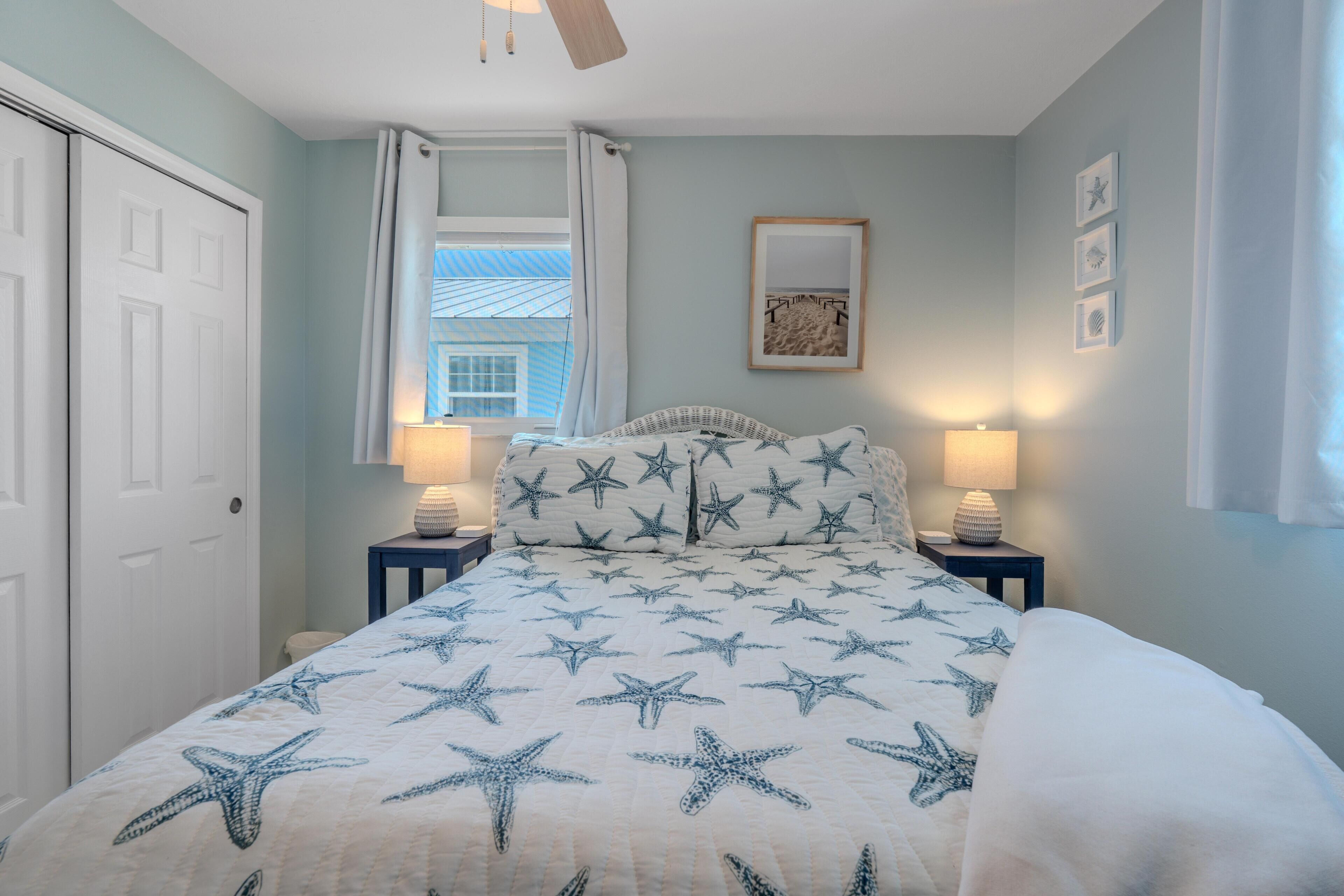 A bedroom with a bed featuring a starfish pattern, two bedside lamps, a ceiling fan, a window with curtains, and a framed picture on the wall.