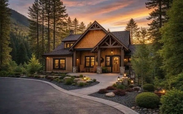 A large, two-story log cabin with a stone foundation, a wrap-around porch, and a driveway leading to the entrance.