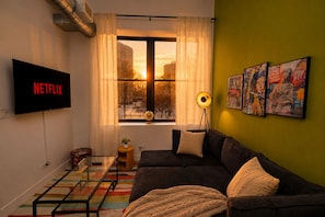 A living room with a sofa, a TV displaying 'NETFLIX,' a window with curtains, a floor lamp, and wall art.