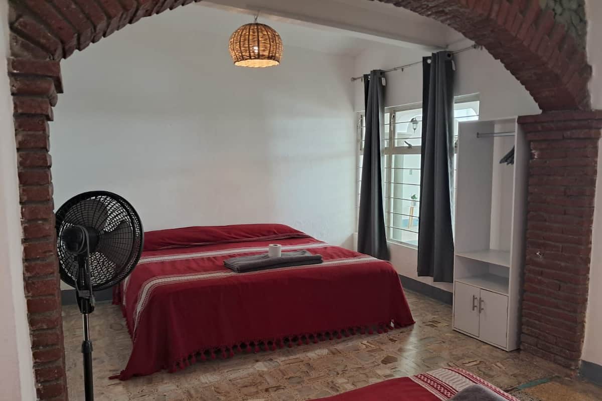 Una habitación con cama, ventilador, armario y ventana con cortinas.