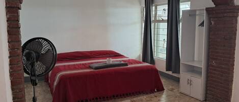 A room with a bed, a standing fan, a wardrobe, and a window with curtains.