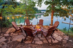 A stone fire pit surrounded by wooden Adirondack chairs, set against a lakeside backdrop with string lights. - Lakefront · Dock · 8 Acres · Hot Tub · 2 Fire Pits (Sevierville)