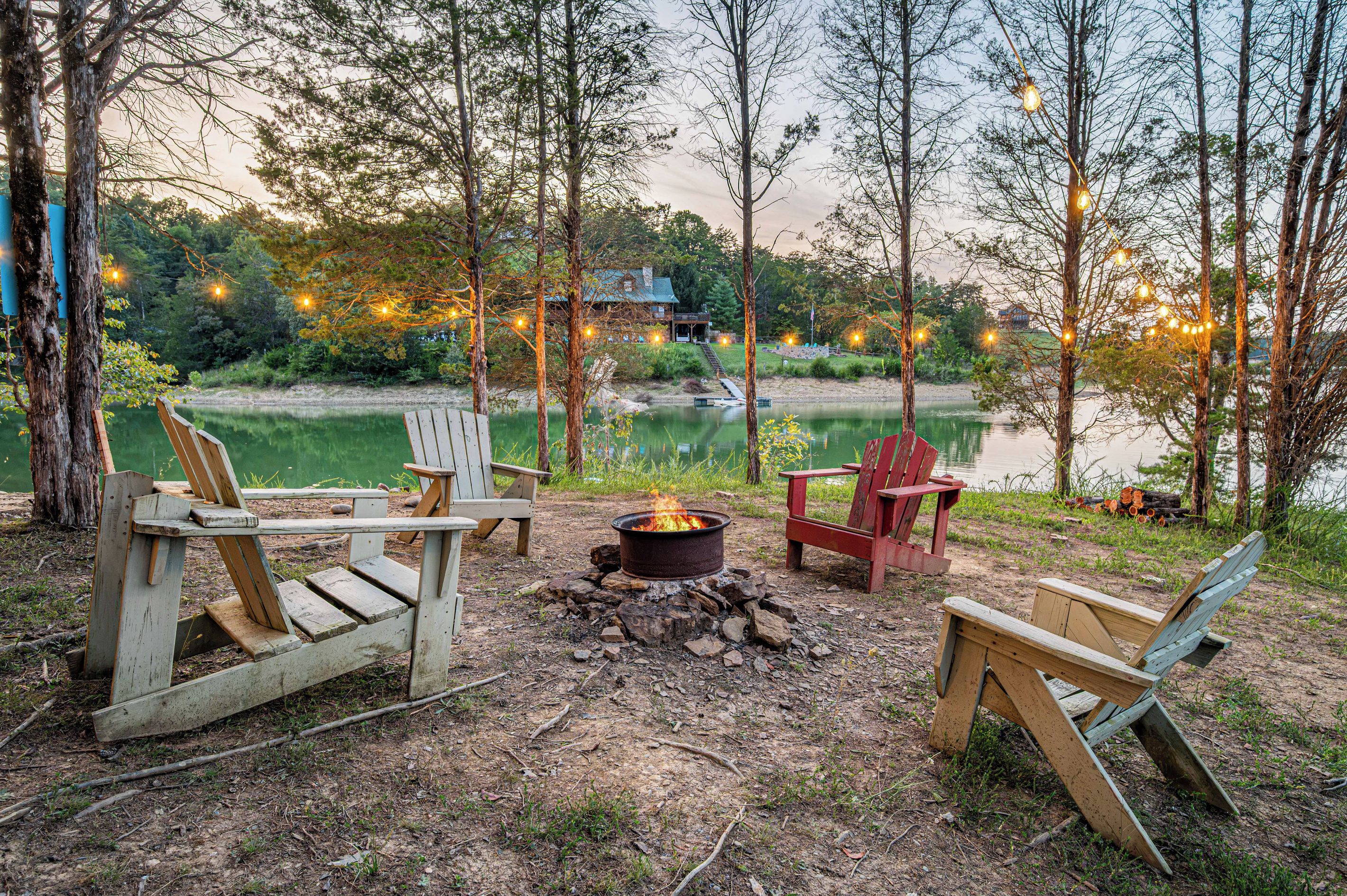 A lakeside campsite with wooden chairs, a fire pit, and string lights.