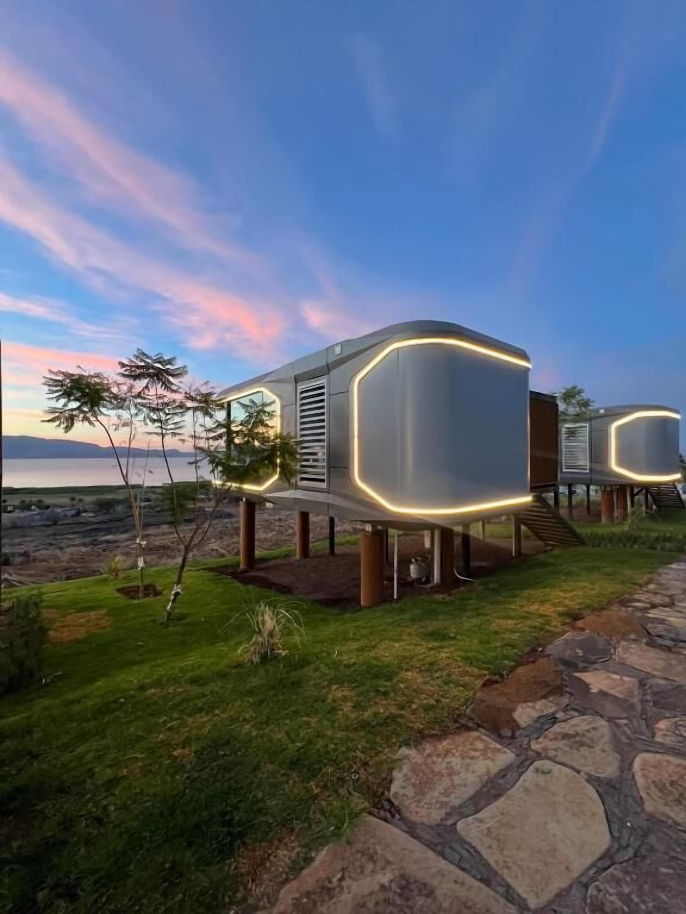 Una estructura moderna de un piso con techo plano, posiblemente una casa móvil o alojamiento temporal, ubicada en un área con césped y un sendero de piedra que conduce hacia ella.
