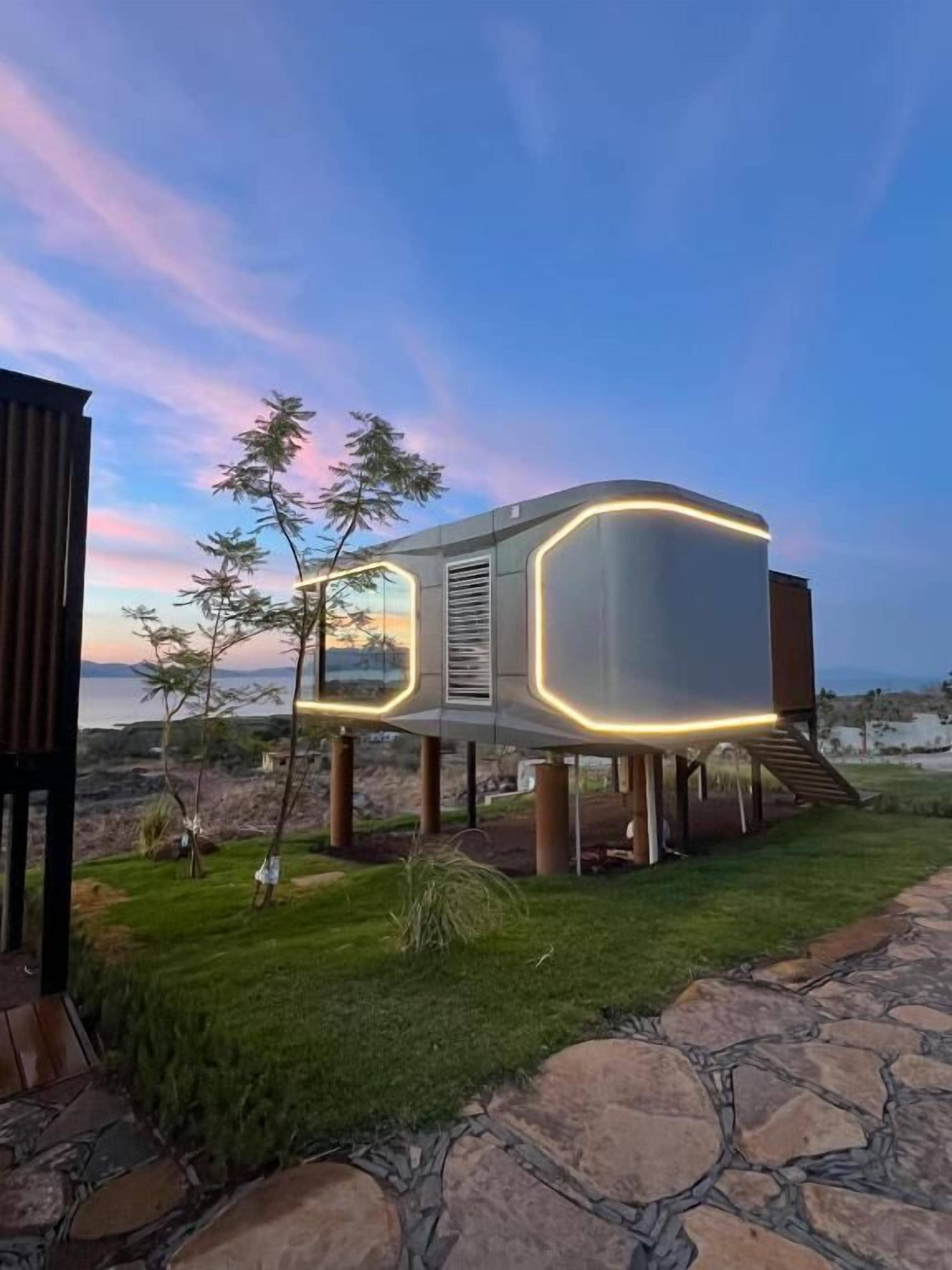 Un edificio moderno con un diseño singular, que presenta una estructura curva y bordes iluminados, ubicado en un área verde con un camino de piedra.