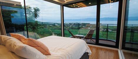 A bedroom with a large bed, a view of the outdoors, and a balcony with chairs.