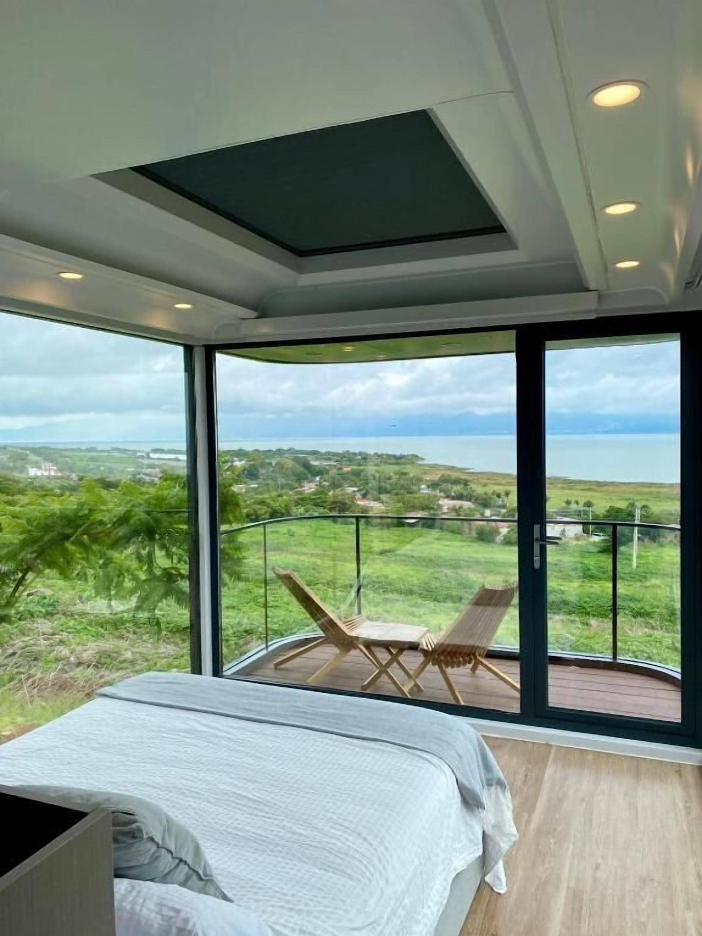 A bedroom with a large window, a bed, and a balcony with a view of a landscape.