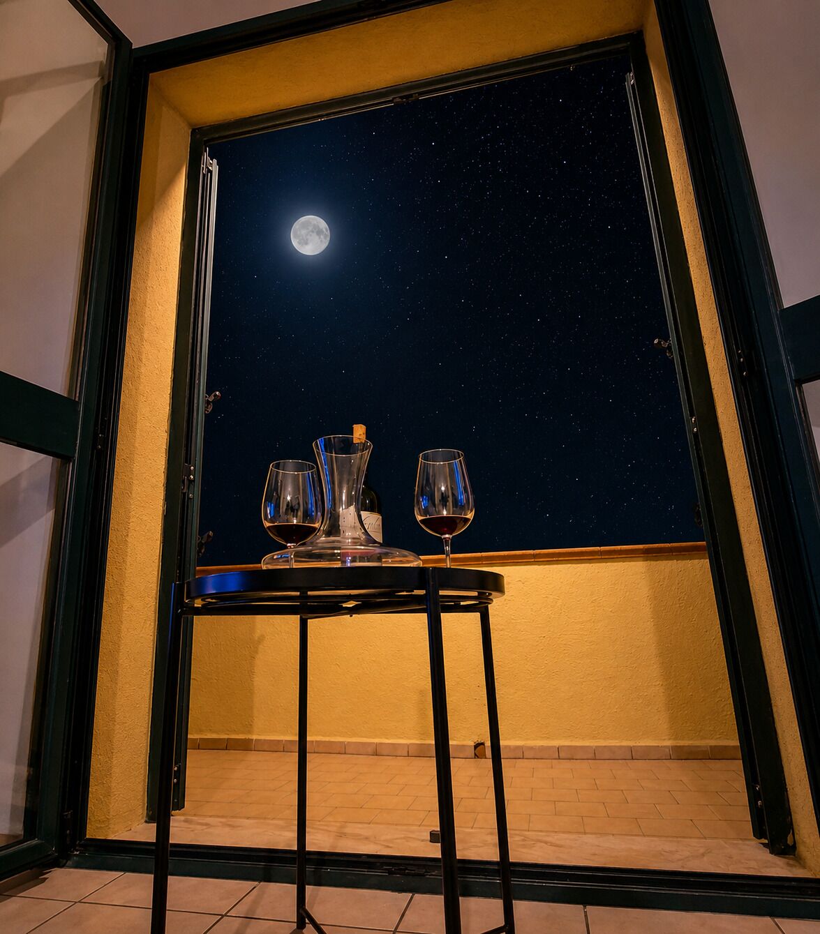 A balcony with a table set for two, a bottle, and a glass of red wine, under a night sky with a visible moon.