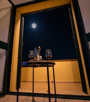 Ein Balkon mit Tisch für Zwei, einer Flasche und einem Glas Rotwein, unter einem Sternenhimmel mit sichtbar erkennbarem Mond.