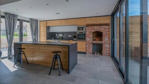 Et moderne køkken med træskabe, en central ø, barstole, en murstenspejs og en stor glasdør.