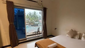 A hotel room with a bed, a ceiling fan, an air conditioning unit, a view of palm trees and a pool outside. - Scarlet Stays (Alibag)