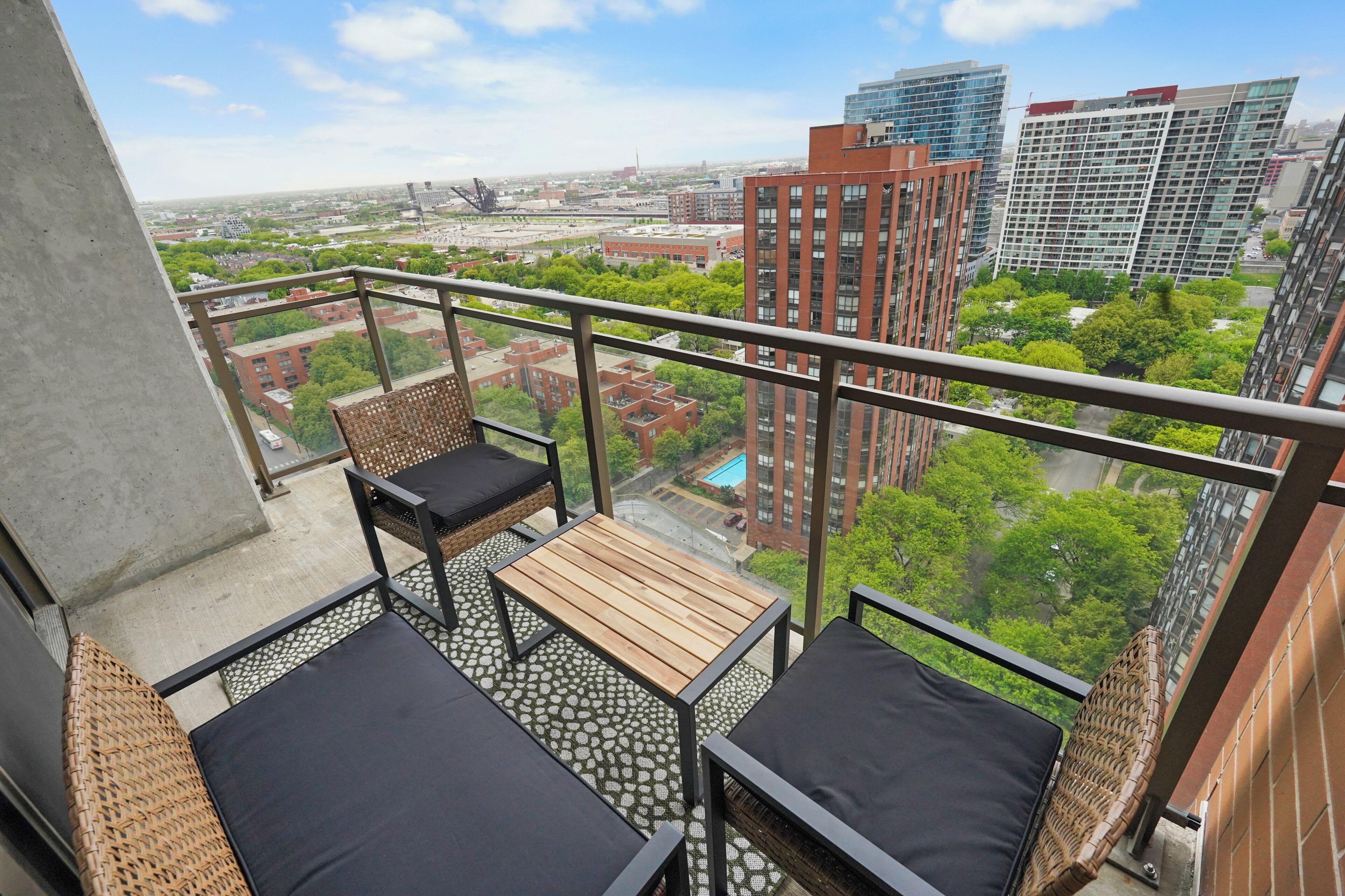 A balcony with wicker furniture, a small wooden table, and a view of a cityscape with buildings and greenery.