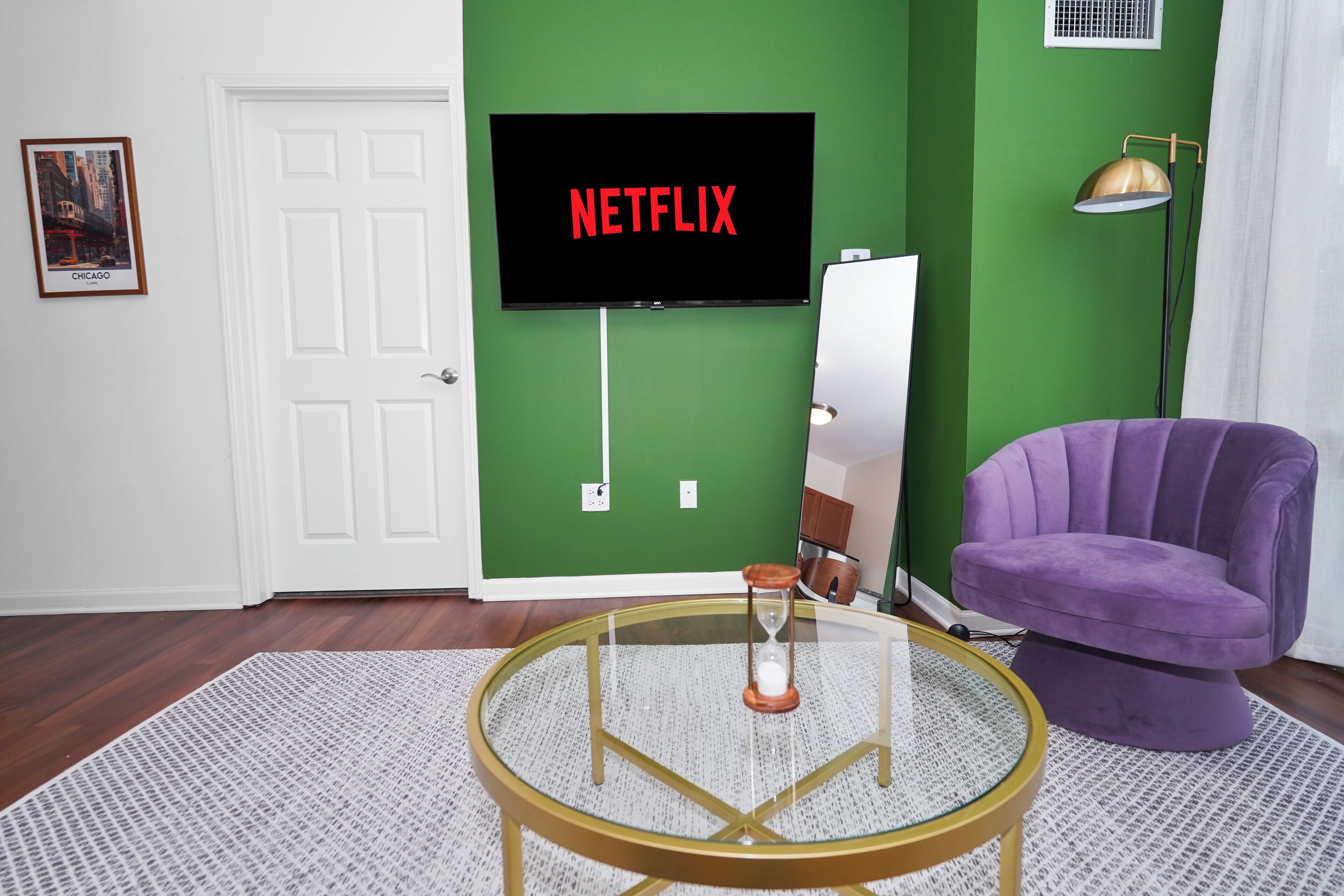 A room with a green wall, a mounted TV displaying 'NETFLIX,' a purple chair, a round glass table with an hourglass, and a white door.