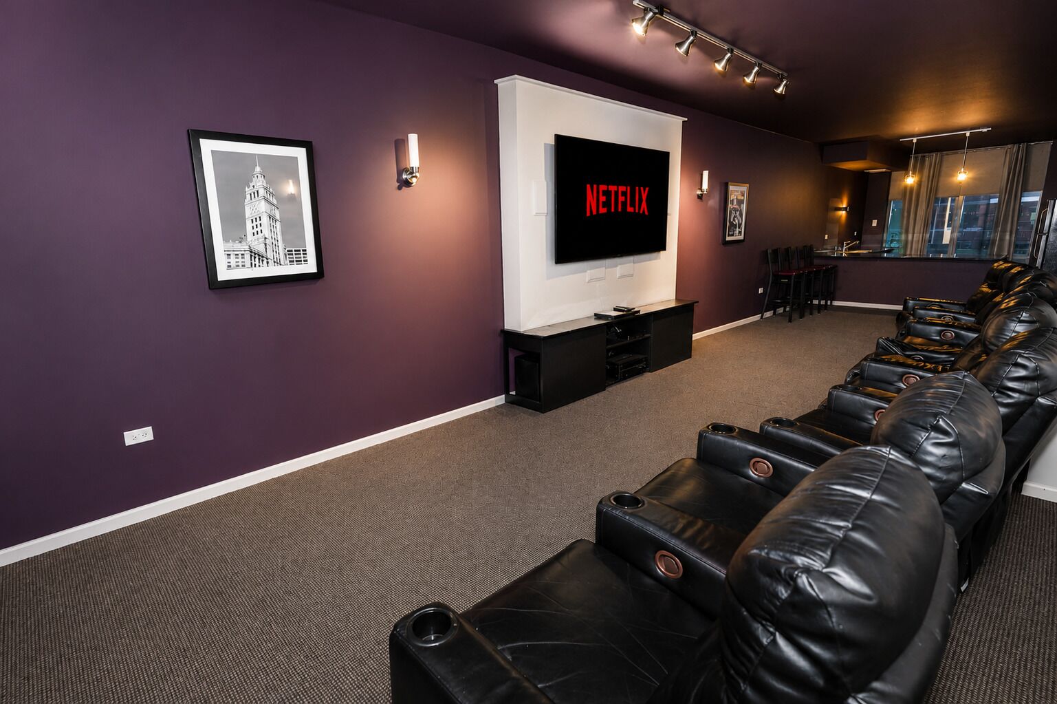 A home theater room with a large TV screen displaying 'NETFLIX,' leather reclining chairs, a framed picture on the wall, and a window with curtains.