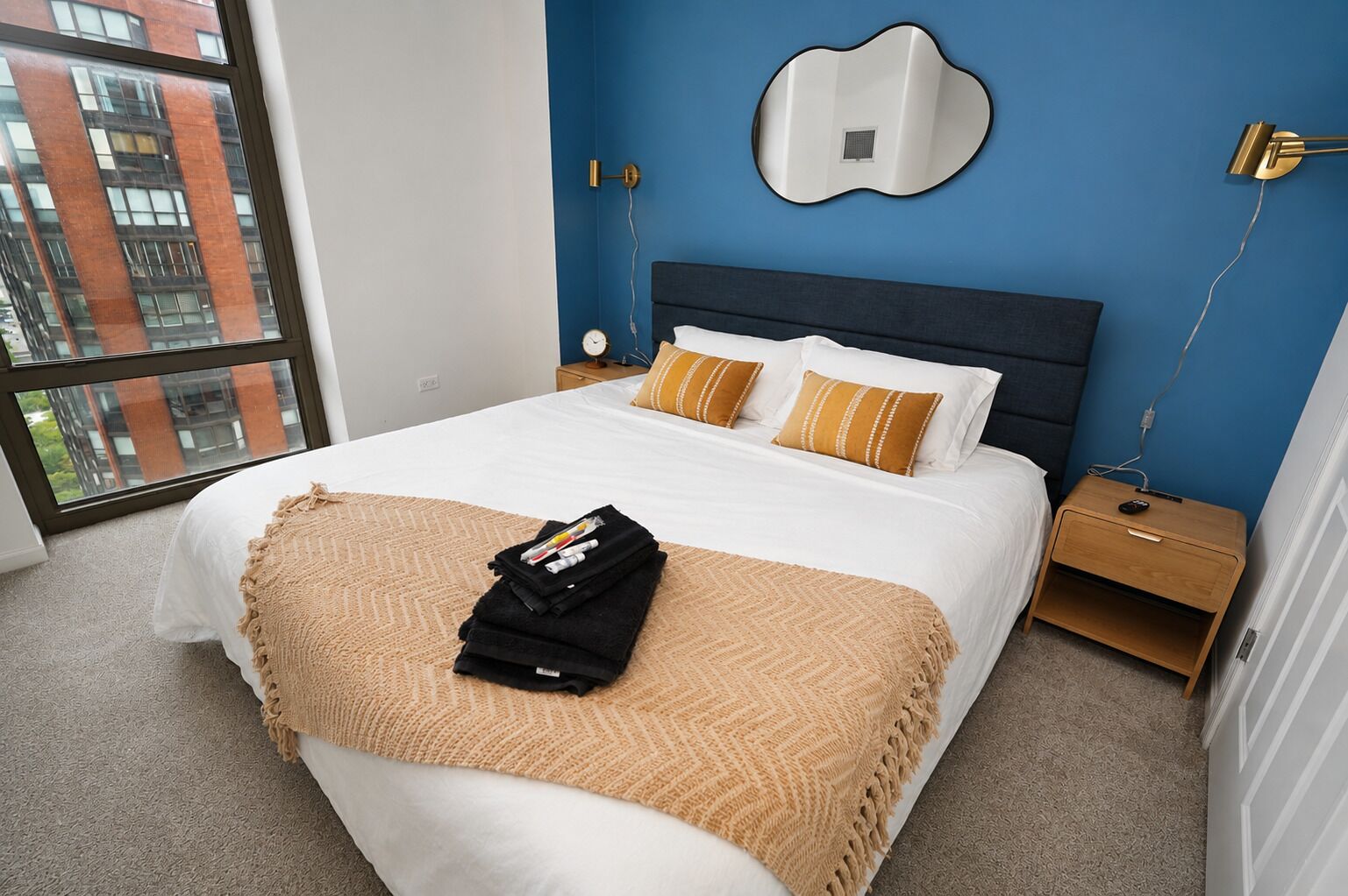 A hotel room with a large bed, a nightstand, a wall-mounted mirror, and a view of a building through a window.