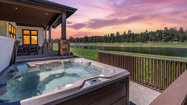 A hot tub on a deck with a view of a lake and trees at sunset. - Brand-New Lakefront Estate | Private Lake + Hot Tub | Outdoor Living (Bend)