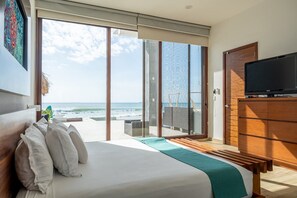 Ein Schlafzimmer mit einem großen Bett, einem Fernseher, einer Holzkommode und einer Glastür, die auf einen Balkon mit Meerblick führt.