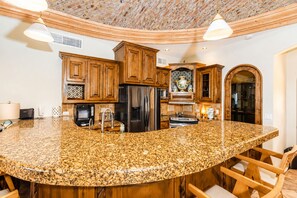 A kitchen with a large granite island, wooden cabinets, stainless steel appliances, and a unique ceiling design.