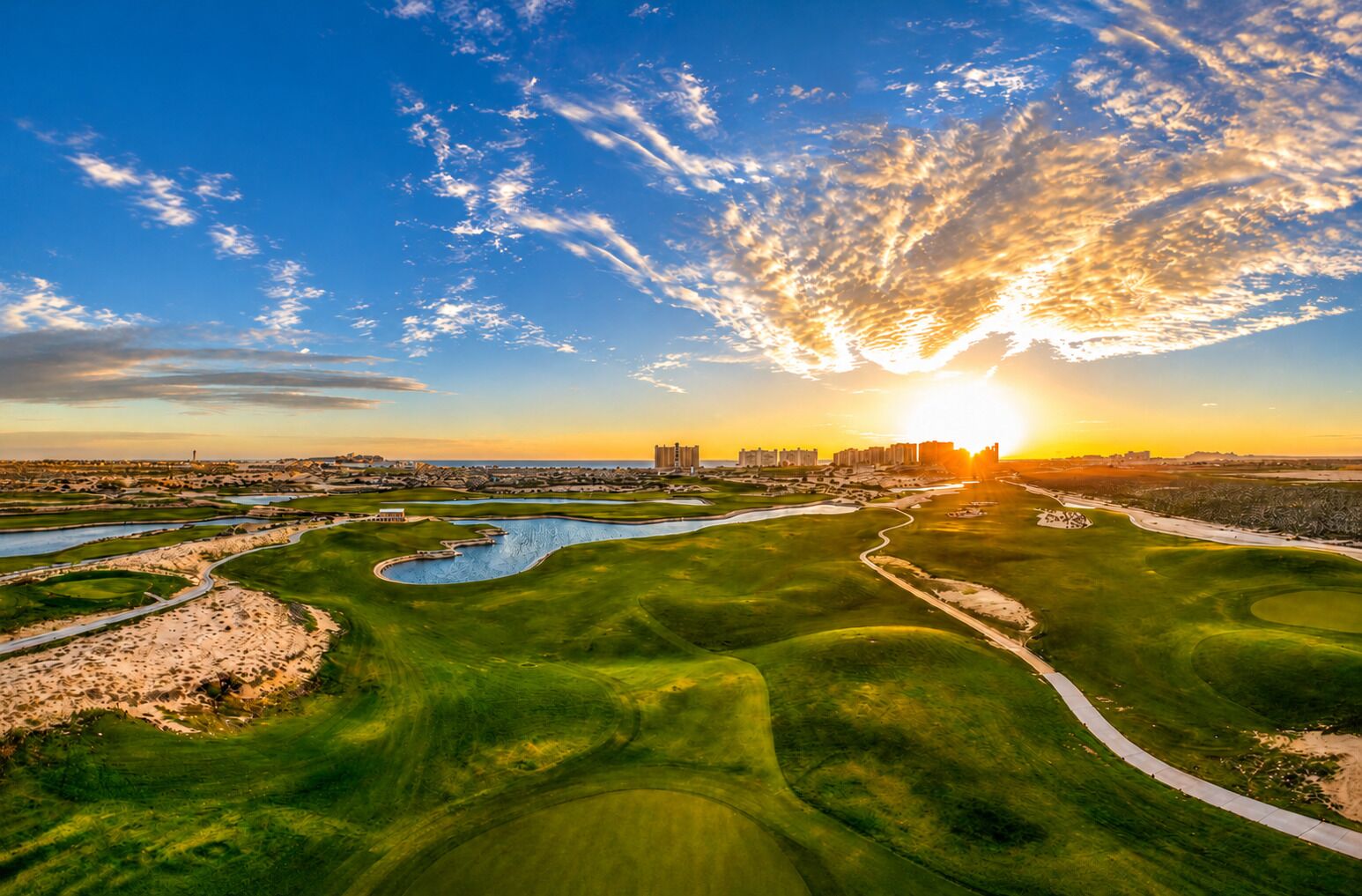 A golf course with a river, a sunset, and buildings in the distance.