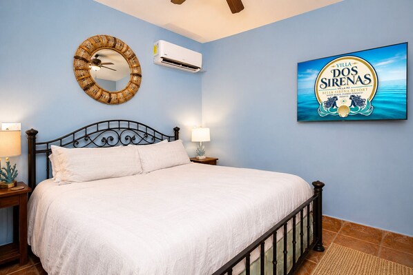 A bedroom with a bed, a wall-mounted air conditioner, a decorative mirror, a nightstand with a lamp, and a framed picture of a resort.