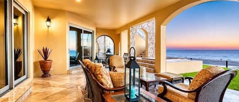 Un patio iluminado por el sol con asientos al aire libre, vista al océano y una puerta de cristal que da al interior.
