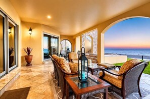 A sunlit patio with outdoor seating, a view of the ocean, and a glass door leading to the interior.